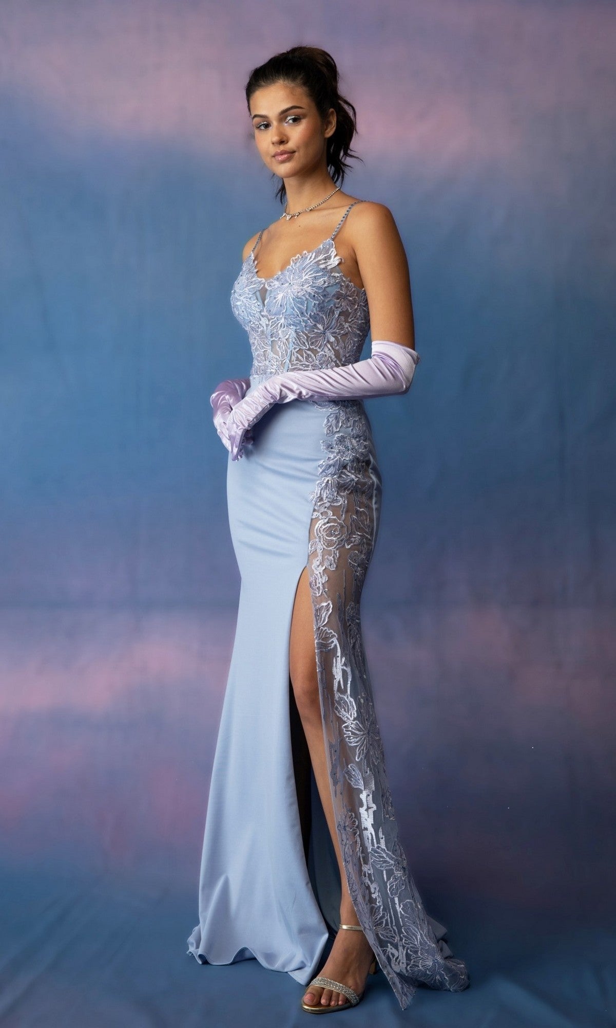 A woman poses against a blue and pink backdrop wearing the Long Prom Dress: Eureka 9889 in Periblue, a sleeveless gown with lace appliques and a thigh-high slit, styled with pink opera gloves and silver sandals.